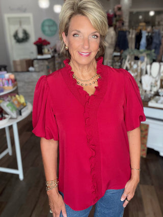 A woman wearing a bright red short-sleeve blouse with ruffle trim and puff sleeves, paired with blue jeans, standing in a boutique setting.
