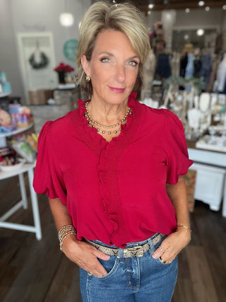 A woman with short blonde hair wearing a bright red ruffled blouse and blue jeans, standing in a boutique setting.