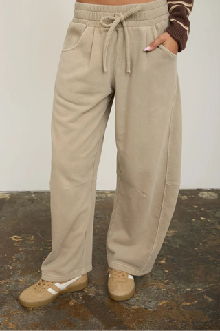 A person wearing loose, wide-leg beige sweatpants with a drawstring waist, paired with a brown and cream striped long-sleeve top and beige sneakers, standing against a plain light wall.