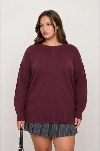A woman modeling an oversized crewneck sweater in a deep burgundy color, paired with a gray pleated skirt, against a plain light gray background.