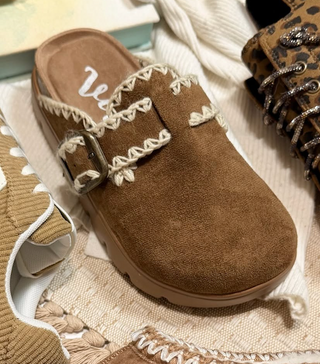 A brown suede mule slipper with cream-colored braided trim and a silver buckle, displayed on a textured beige knit blanket alongside other shoes.