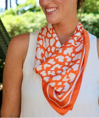 A woman wearing a white sleeveless top and a bright orange and white patterned scarf, photographed outdoors with green foliage and a tree trunk in the background.