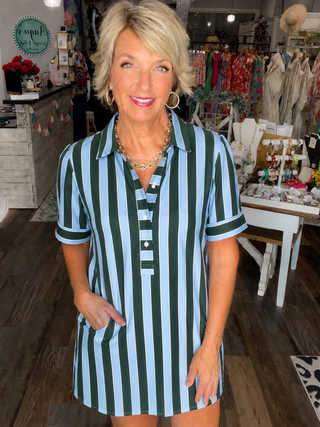 A woman modeling a short-sleeve collared shirt dress with vertical light blue and dark green stripes, standing inside a retail boutique.