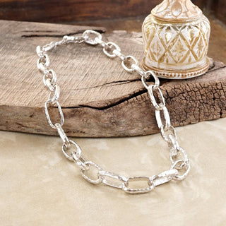 A silver chain link necklace with large, polished oval links, displayed on a rustic wooden block next to a decorative ceramic container.