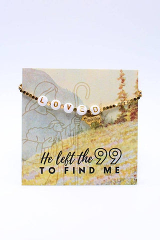 A gold beaded bracelet with white letter beads spelling 'LOVED' and a small gold lamb charm, displayed on a printed card that reads 'He left the 99 TO FIND ME'.