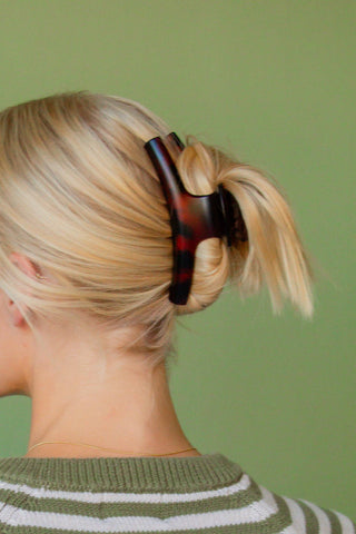 A close-up back view of a person with blonde hair tied up in a low ponytail using a large tortoiseshell hair claw clip, set against a solid sage green background.