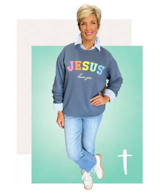 Woman wearing a blue sweatshirt with multicolor JESUS loves you graphic, paired with light wash jeans and white sneakers on teal background.