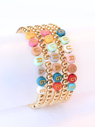 A stack of five beaded bracelets on a white wristband, featuring gold-plated round beads and pastel letter beads spelling 'COLORAGE' in multiple colors.