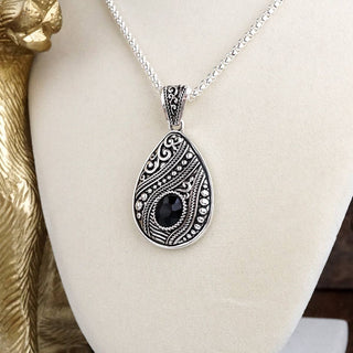 A silver teardrop pendant necklace with intricate filigree cutouts and a central black stone, displayed on a cream-colored jewelry bust.