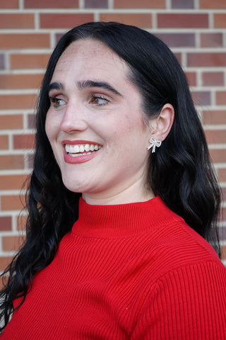 Woman wearing a red sweater  and white bow earrings in front of a brick wall