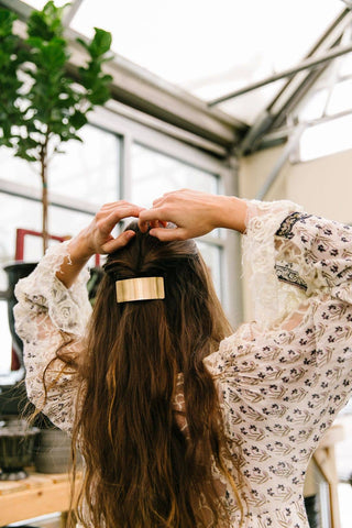 A woman with long, wavy brown hair seen from behind, wearing a cream lace top and adjusting her hair with a wide, rectangular gold barrette in a bright indoor setting with plants and windows.