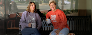 Two women sitting on a bench wearing Small Town Proud sweatshirts, holding coffee mugs