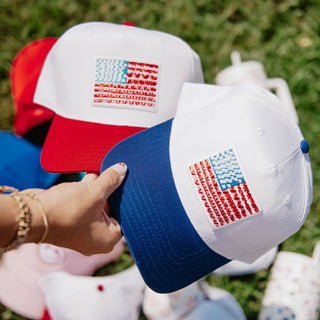 Two baseball caps with American flag designs held by a hand against a blurred grass background.