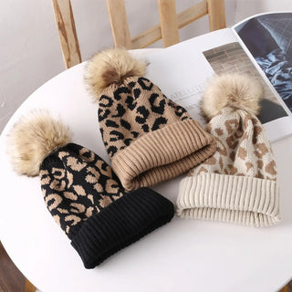Three leopard print beanies with pom-poms on a white surface.