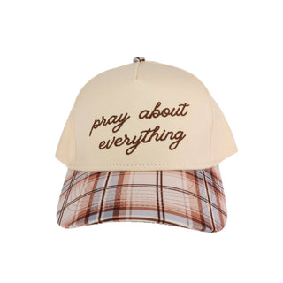 A baseball cap with a cream-colored crown and brown embroidered text reading 'pray about everything' on the front, paired with a brown, beige, and white plaid visor.