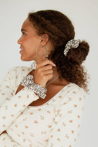 A woman shown in profile wearing a leopard-print scrunchie in her hair and a matching leopard-print wristband, paired with a white floral long-sleeve top against a plain light background.