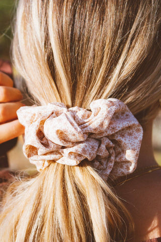 A close-up shot of a blonde scrunchie with a light beige and white floral pattern, tied around a person's low ponytail.