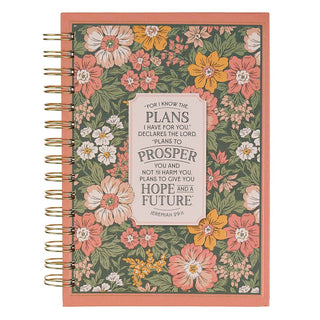 A spiral-bound planner or notebook with a coral pink border and a floral patterned cover featuring pink, yellow, and white flowers on a green background. A central cream label displays a Bible verse from Jeremiah 29:11.