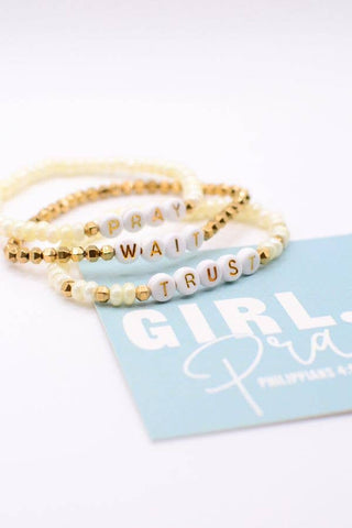 Three beaded stretch bracelets with gold and white beads, featuring lettered beads spelling out 'PRAY WAIT TRUST' and gold spacer beads, displayed on a light blue card.