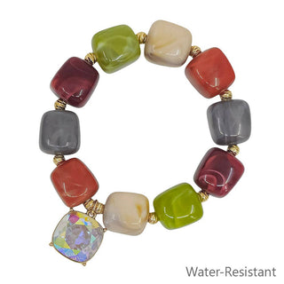 A multicolor beaded stretch bracelet with rounded square glass beads in green, red, gray, cream, and burgundy, separated by gold-toned spacers, featuring a large iridescent rhinestone charm at the bottom. The text 'Water-Resistant' is printed in the bottom right corner.
