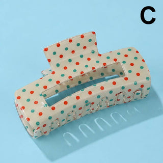 A large claw hair clip with a cream base and red and teal polka dots, shown on a light blue background with a black letter 'C' in the top right corner.