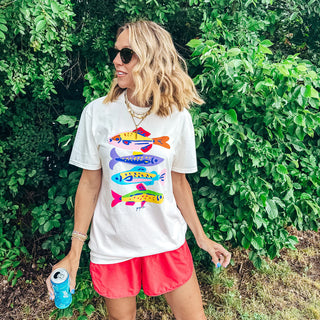 Woman wearing a colorful graphic t-shirt and pink shorts standing in front of green foliage.