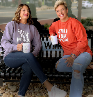 Two women sitting on a bench wearing Small Town Proud sweatshirts, holding coffee mugs