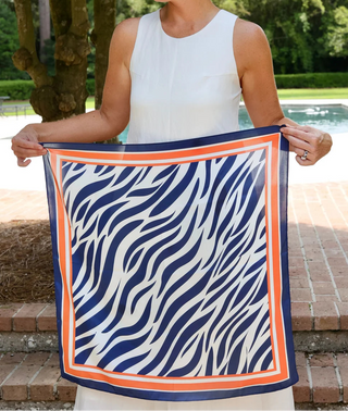 A woman outdoors by a pool holds up a square scarf with a navy blue and white zebra-like pattern and a bright orange border.