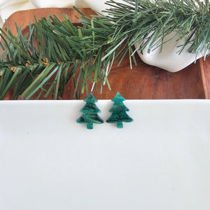 Green tree-shaped earrings on a white surface with a wooden board and greenery in the background.