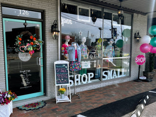 Boutique storefront decorated with balloons, wreath, chalkboard sale sign, and mannequins in colorful outfits displayed in the window with shop small signage.