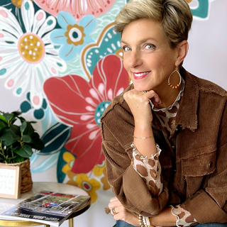Woman wearing a brown corduroy jacket with patterned blouse and hoop earrings, seated in front of a colorful floral mural.