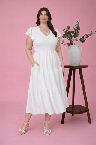 Woman in a white dress standing next to a wooden side table with a plant on a pink background