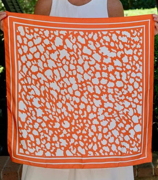 A woman holding up a square orange scarf with a white abstract animal-like spot pattern and a white border, set against a background of green foliage and a porch.